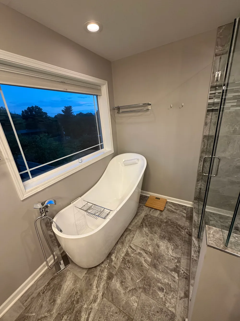 A freestanding white bathtub beneath a wide window beside a tiled shower.