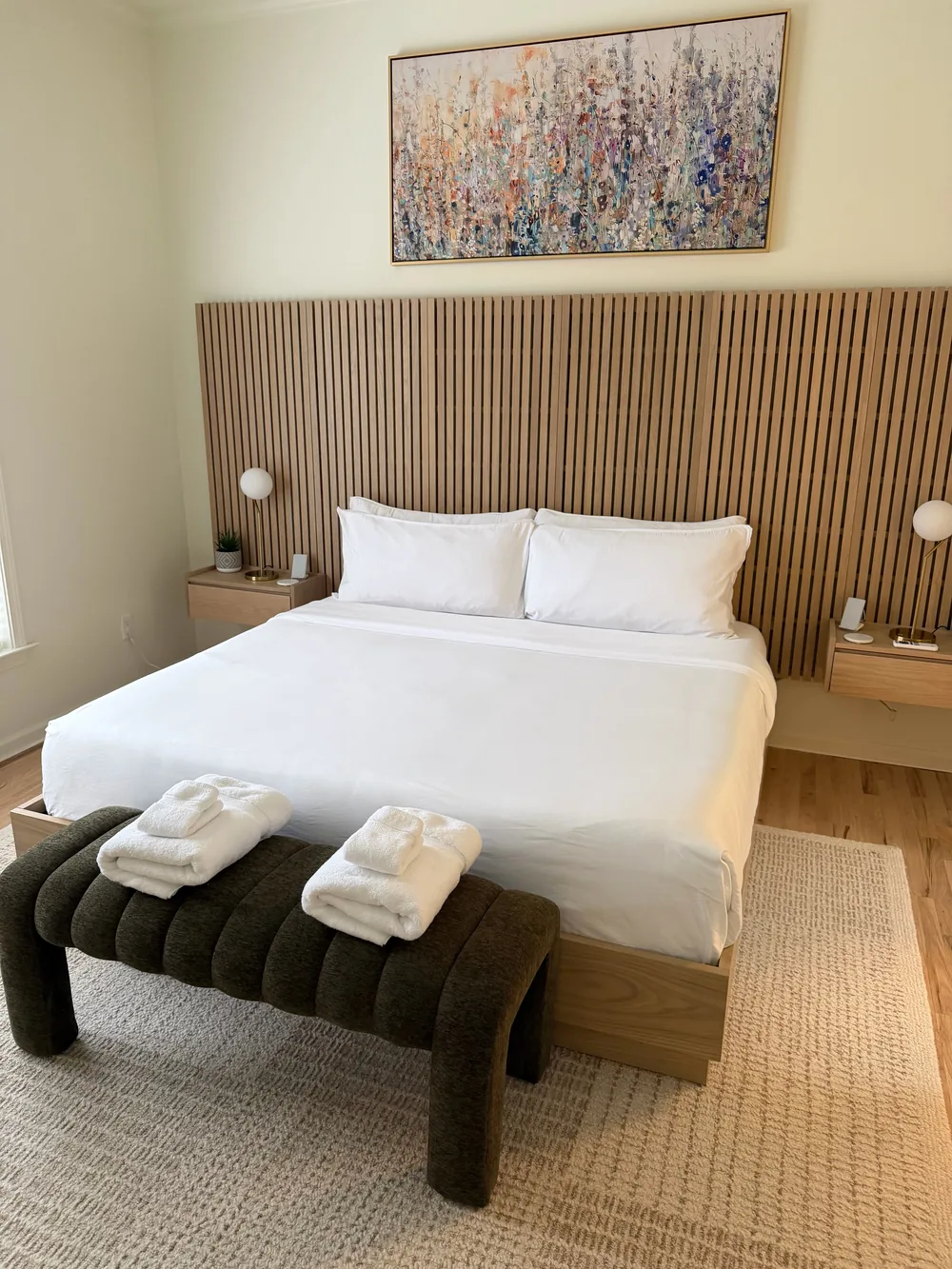 White bedding, a slatted wood headboard, and folded towels on an upholstered bench.